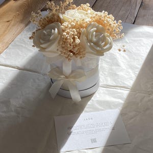 Peut inclure: Une boîte blanche avec un ruban blanc et un bouquet de fleurs de cire blanches. La boîte porte une étiquette qui indique "JADE" et comprend le texte "CHAQUE BOUQUET EST REALISE A LA MAIN A PARTIR DE CIRE DE SOJA NATURELLE, DANS LE RESPECT DE L'ARTISANAT ET DU DETAIL. J'ESPERE QU'IL ILLUMINERA VOTRE INTERIEUR ET APPORTERA UNE TOUCHE D'ELEGANCE A VOTRE QUOTIDIEN."
