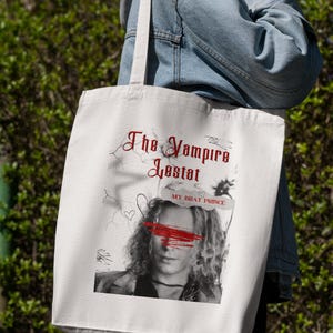 May include: White tote bag featuring a black and white image of a person with red paint over their eyes. The text "The Vampire Lestat" and "My Brat Prince" is printed on the bag.