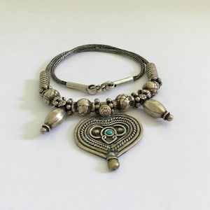 Peut inclure: Un collier en argent avec un pendentif en forme de cœur orné de motifs complexes et d'une pierre turquoise. Le collier a une chaîne avec de petites perles en argent.