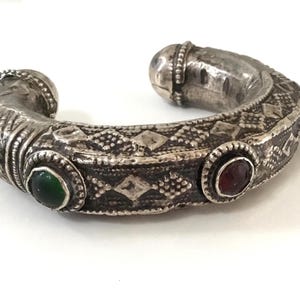 Op de afbeelding: Een zilverkleurige manchetarmband met een ingewikkeld, in reliëf aangebracht geometrisch patroon over de band. Het sieraad is voorzien van een ronde groene steen en een ronde rode steen, gezet in decoratieve metalen randen aan de voorzijde.