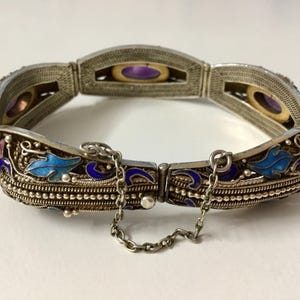 May include: A silver bracelet with blue and purple enamel detailing and a chain closure. The bracelet features a floral design and is decorated with small, round stones.