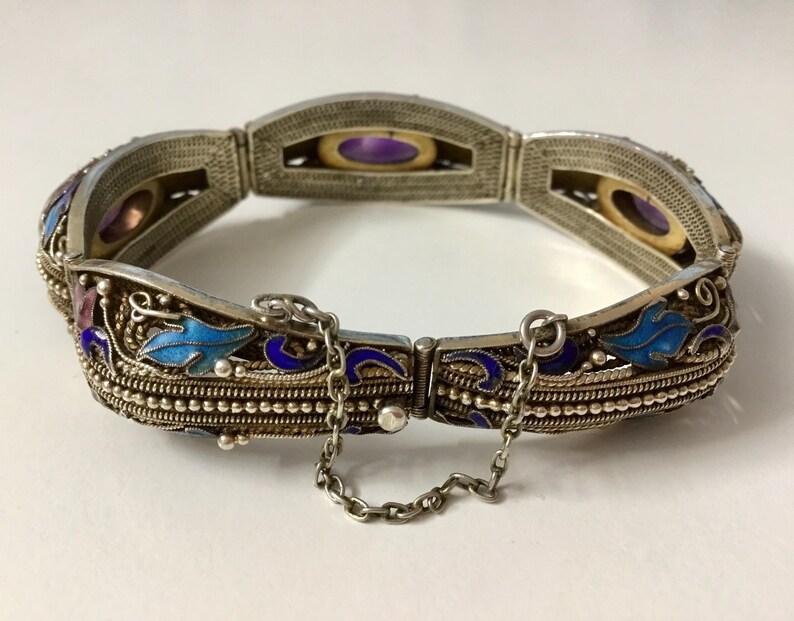 May include: A silver bracelet with blue and purple enamel detailing and a chain closure. The bracelet features a floral design and is decorated with small, round stones.