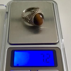 May include: A silver ring with an oval-shaped tiger's eye stone set in a filigree design. The ring is on a digital scale that reads 7.12 grams.