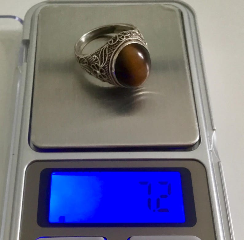 May include: A silver ring with an oval-shaped tiger's eye stone set in a filigree design. The ring is on a digital scale that reads 7.12 grams.
