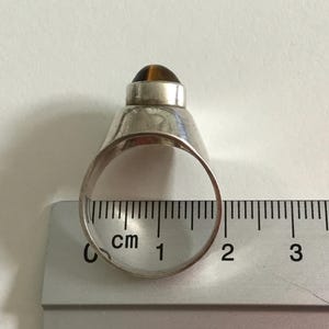 May include: Silver ring with a brown tiger's eye stone set in a dome shape. The ring is measured on a ruler with centimeters.