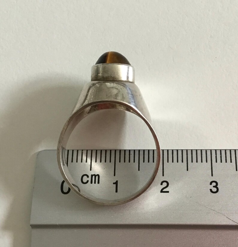 May include: Silver ring with a brown tiger's eye stone set in a dome shape. The ring is measured on a ruler with centimeters.