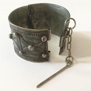 Op de afbeelding: Een brede, zilverkleurige metalen manchetarmband met een ingewikkeld reliëfpatroon, verhoogde bolvormige noppen en een scharnierende opening. Een dunne metalen ketting is aan één kant bevestigd en verbonden met een lange, slanke metalen pin.