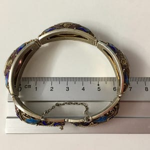 May include: A silver bracelet with blue and purple enamel and filigree detailing. The bracelet is 6 cm in length and has a chain closure.