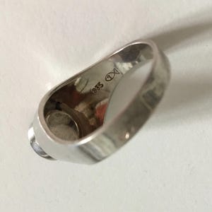 May include: Silver ring with a round, white stone set in the center. The inside of the band is marked with "935" and a symbol.