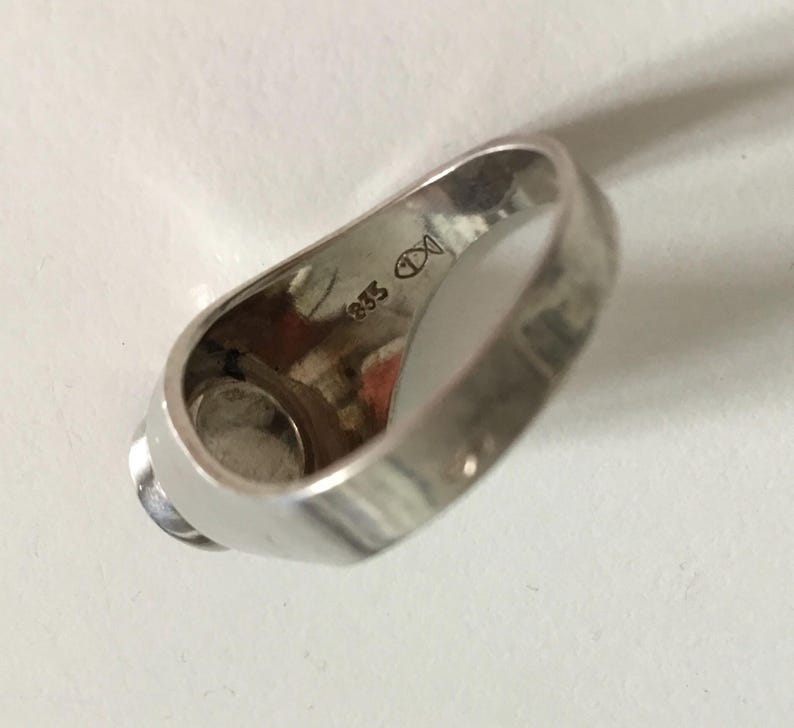 May include: Silver ring with a round, white stone set in the center. The inside of the band is marked with "935" and a symbol.