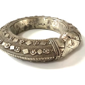 Op de afbeelding: Een zilverkleurige, sierlijke armband. De armband heeft gedetailleerde patronen van ruiten, bloemen en touwachtige texturen. Het metaal heeft een licht verouderde uitstraling, met zichtbare slijtage, wat suggereert dat het vintage of antiek is.