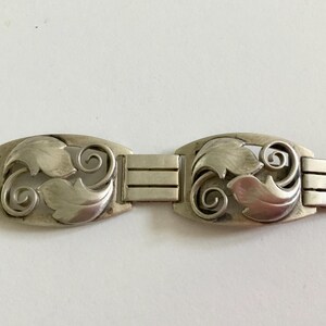 May include: A silver bracelet with a repeating pattern of stylized leaves and swirls. The bracelet is made of linked sections, each with a decorative element.