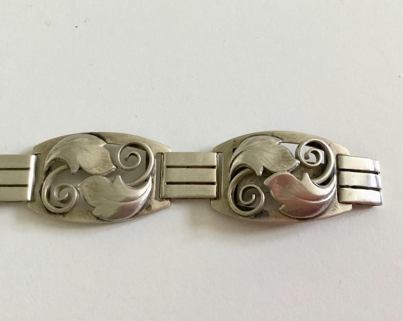 May include: A silver bracelet with a repeating pattern of stylized leaves and swirls. The bracelet is made of linked sections, each with a decorative element.