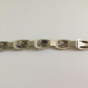 May include: A silver bracelet with a repeating pattern of rectangular links featuring a stylized floral design.