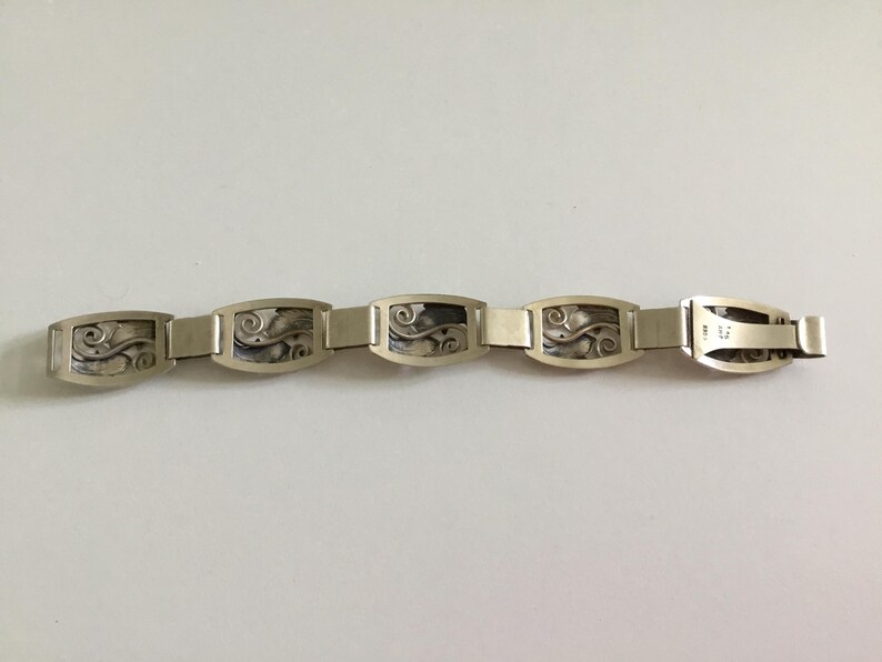 May include: A silver bracelet with a repeating pattern of rectangular links featuring a stylized floral design.