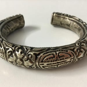 Puede incluir: Un brazalete de plata con intrincados diseños florales y geométricos. El brazalete está abierto y tiene una forma ligeramente curvada.