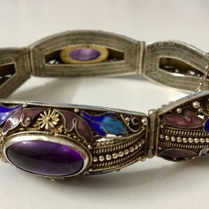 May include: A silver bracelet with an intricate design featuring purple, blue, and white enamel. The bracelet has a hinged clasp and is decorated with a large amethyst gemstone.