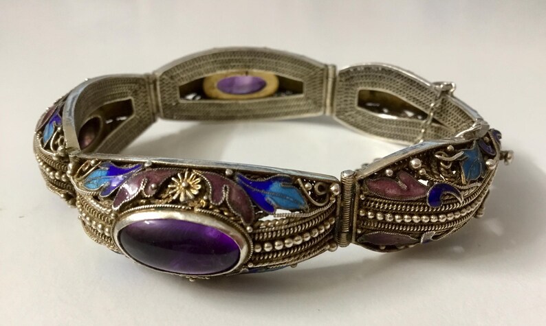 May include: A silver bracelet with an intricate design featuring purple, blue, and white enamel. The bracelet has a hinged clasp and is decorated with a large amethyst gemstone.