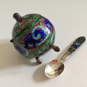 Puede incluir: Un azucarero en forma de globo de plata y esmalte con una cuchara a juego. El bol está decorado con esmalte azul, verde y rosa y diseños intrincados. La cuchara también está decorada con esmalte.