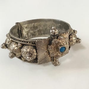 Op de afbeelding: Een zilverkleurige scharnierende armband met een getextureerd oppervlak en meerdere afgeronde, koepelvormige uitsteeksels. Het centrale sluitingsmechanisme is vierkant en voorzien van een kleine, ronde, felblauwe steen als accent.