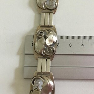 May include: A silver bracelet with a repeating pattern of stylized leaves. The bracelet is measured against a ruler, showing the length of each link is approximately 1 inch.