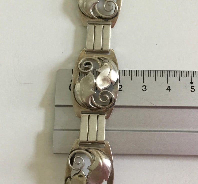 May include: A silver bracelet with a repeating pattern of stylized leaves. The bracelet is measured against a ruler, showing the length of each link is approximately 1 inch.