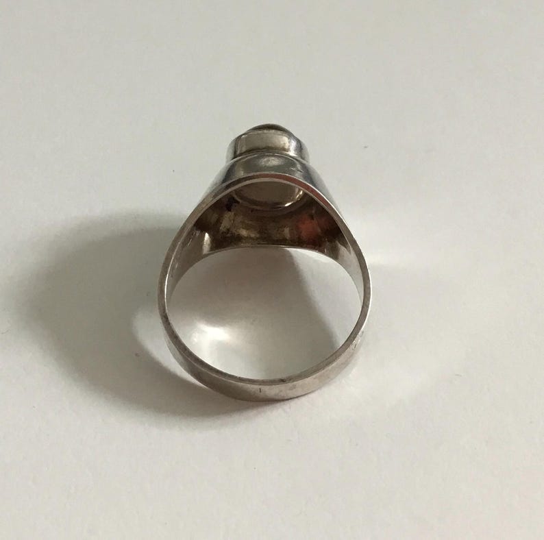 May include: A silver ring with a round, opaque white stone set in the center. The ring has a wide band and a slightly domed top.