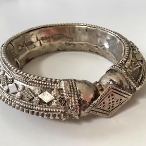 Op de afbeelding: Een zilverkleurige armband met ingewikkelde details. De armband heeft geometrische ruitvormen, kralenaccenten en een getextureerd oppervlak. Het ontwerp suggereert een vintage of etnische sieradenstijl.