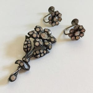 Op de afbeelding: Een zilveren hanger in de vorm van een bloem met een hangende druppel en twee bijpassende oorringen. De sieraden zijn versierd met kleine, heldere strass steentjes.