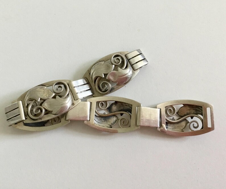 May include: A silver bracelet with a delicate, intricate design of leaves and swirls. The bracelet is made of multiple linked sections, each featuring a unique pattern.