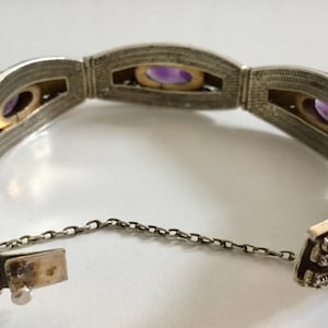 May include: A silver bracelet with a chain closure and intricate filigree detailing. The bracelet features four oval-shaped amethyst stones set in the center of each section. The bracelet is decorated with blue and white enamel.