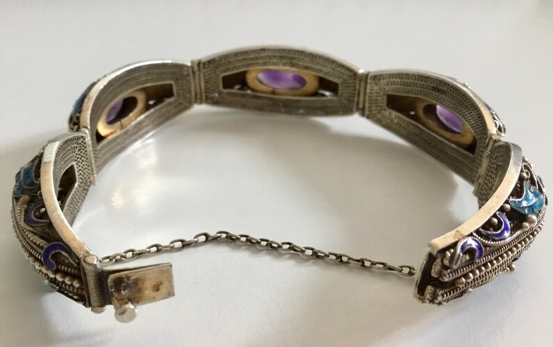 May include: A silver bracelet with a chain closure and intricate filigree detailing. The bracelet features four oval-shaped amethyst stones set in the center of each section. The bracelet is decorated with blue and white enamel.
