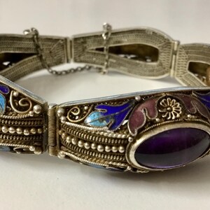 May include: A silver bracelet with an intricate design featuring enamel flowers and leaves in shades of blue, purple, and pink. The bracelet has two oval-shaped amethyst stones.