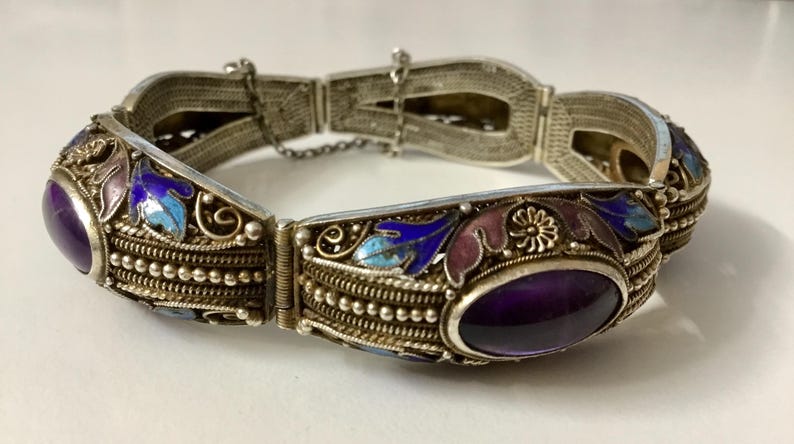 May include: A silver bracelet with an intricate design featuring enamel flowers and leaves in shades of blue, purple, and pink. The bracelet has two oval-shaped amethyst stones.