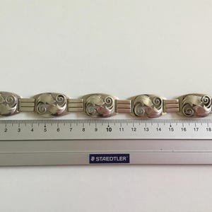 May include: A silver bracelet with a repeating pattern of stylized leaves. The bracelet is approximately 19 centimeters long.