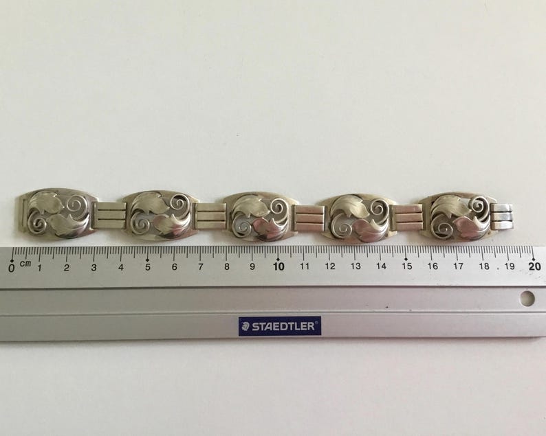 May include: A silver bracelet with a repeating pattern of stylized leaves. The bracelet is approximately 19 centimeters long.