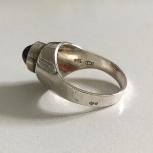 May include: A silver ring with a black stone set in a raised, geometric design. The ring is marked with "925" and a fish symbol.