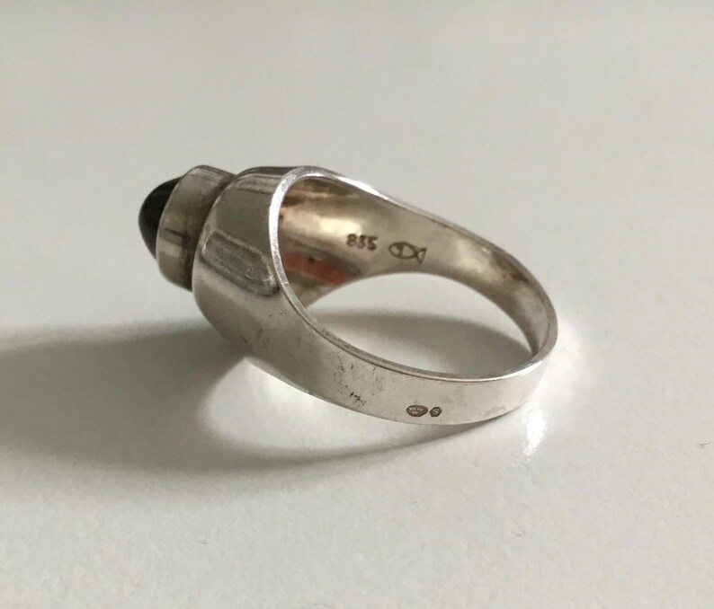 May include: A silver ring with a black stone set in a raised, geometric design. The ring is marked with "925" and a fish symbol.