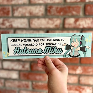 May include: A teal and white bumper sticker with the text "KEEP HONKING! I'M LISTENING TO GLOBAL VOCALOID POP SENSATION Hatsune Miku." Features a cartoon illustration of Hatsune Miku. The sticker is held by a hand.