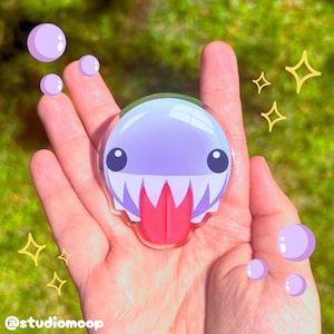 May include: A circular acrylic charm featuring a light purple creature with a wide mouth, sharp white teeth, and a bright pink tongue. The design includes small dark eyes and is surrounded by graphic illustrations of purple bubbles and yellow stars.