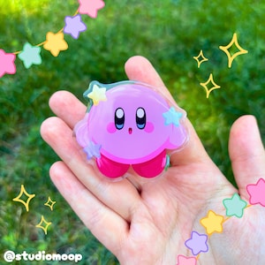 Puede incluir: Un pin de acrílico transparente que muestra al personaje redondo y rosa Kirby con grandes ojos azules y negros, una pequeña boca abierta y pies de color rosa brillante. El diseño incluye pequeñas estrellas claras alrededor, con el texto @studiomoop.