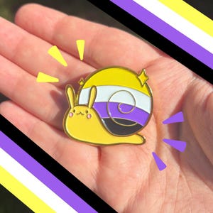 May include: A gold-toned metal enamel pin shaped like a snail with a yellow body and a shell featuring horizontal stripes of yellow, white, purple, and black. The snail has small pink cheeks and a simple face, accented by small star-shaped sparkles.