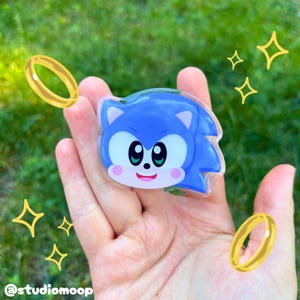 Könnte beinhalten: Eine Anstecknadel mit dem Kopf der Figur Sonic the Hedgehog, mit großen schwarzen Augen, einer kleinen schwarzen Nase und rosa Wangen. Das Design enthält goldene Ringgrafiken und Sternsymbole. Der Text @studiomoop ist unten aufgedruckt.
