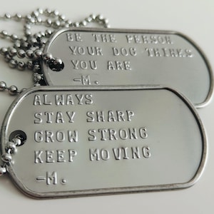 Pode incluir: Duas coleiras com placas de identificação prateadas. Uma placa diz "BE THE PERSON YOUR DOG THINKS YOU ARE -M." A outra placa diz "ALWAYS STAY SHARP GROW STRONG KEEP MOVING -M."