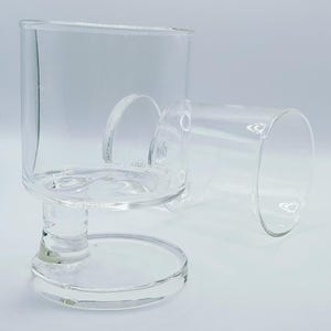 May include: Two clear glass candle holders. One is upright, with a square top and a circular base. The other is lying on its side. The candle holders are made of transparent glass and are designed to hold candles.