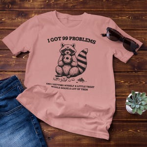 以下が含まれることがあります： ピンクのTシャツに、ドーナツを持ったアライグマの白黒イラストが描かれています。Tシャツには、「99の問題を抱えていて、ちょっとしたご褒美を自分に与えれば、多くの問題が解決するだろう」という文字が書かれています。
