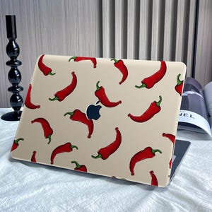 Puede incluir: Una funda para portátil con un patrón repetido de chiles rojos con tallos verdes sobre un fondo beige. La funda está diseñada para adaptarse a un portátil, con el logotipo de Apple visible. El diseño es una forma divertida y única de personalizar un portátil.