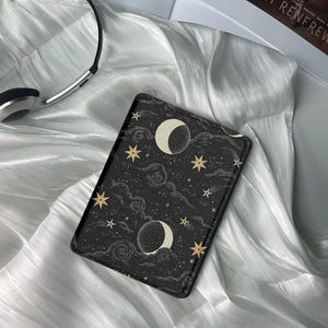 May include: A black e-reader case with a celestial pattern featuring gold stars, moons, and clouds. The case has a textured surface.