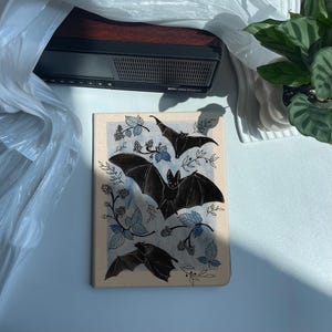 May include: A beige canvas print featuring a blue and black illustration of bats flying over a blackberry bush. The bats are black and the blackberry bush is blue with black berries.