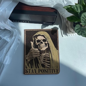 Könnte beinhalten: Ein braunes und schwarzes gerahmtes Poster mit einem Skelett, das eine Kapuzenrobe trägt und einen Daumen nach oben zeigt. Der Text auf dem Poster lautet "Stay Positive".